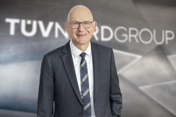 Ein lächelnder Mann mit Glatze und Brille trägt einen dunkelblauen Anzug, ein weißes Hemd und eine gemusterte Krawatte. Er steht vor einer modernen, metallischen Wand mit dem Schriftzug ‚TÜV NORD GROUP‘